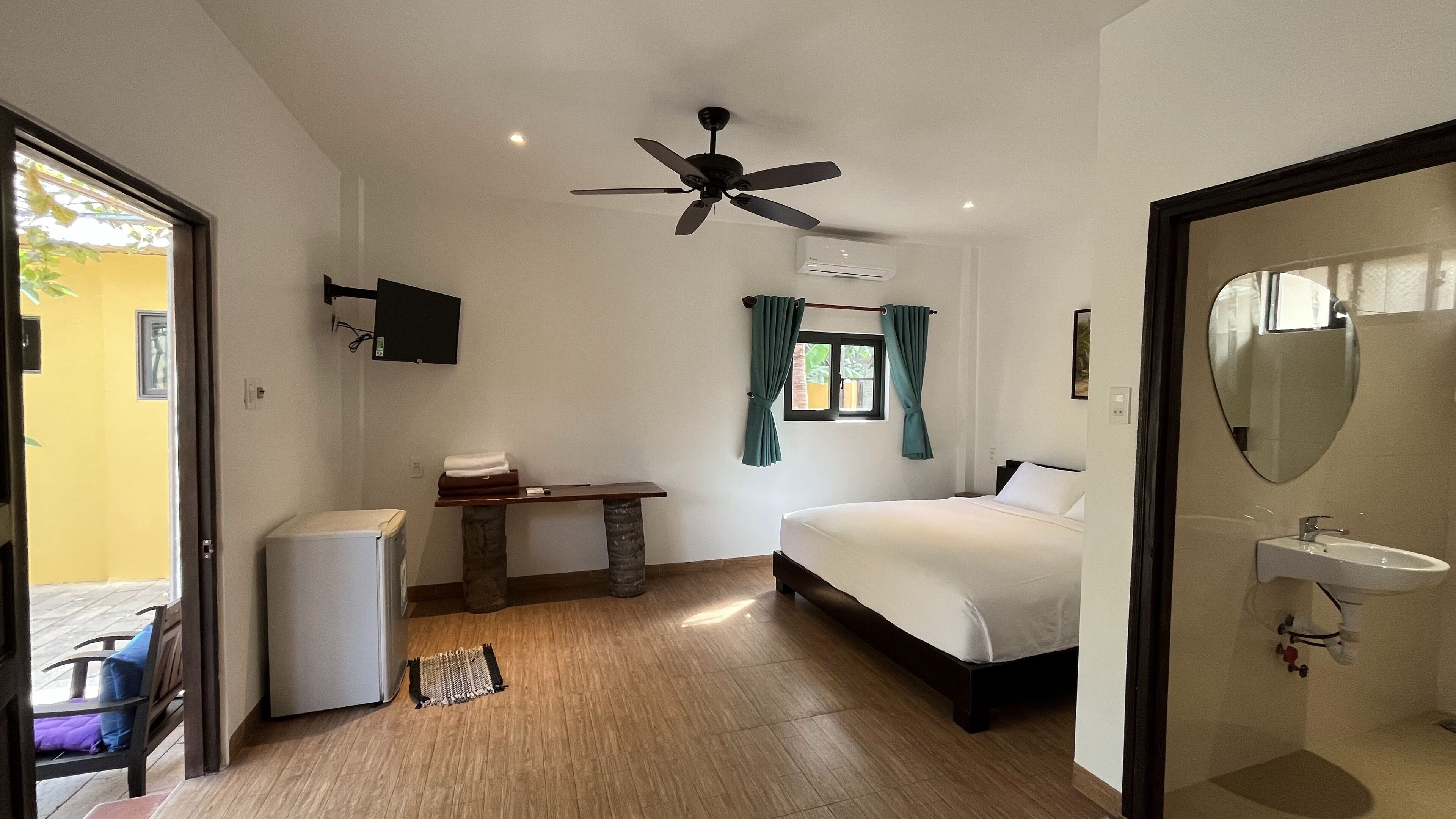 Quarto com cama, escrivaninha, ventoinha no teto, televisão, janela com cortinas e banheiro com pia.