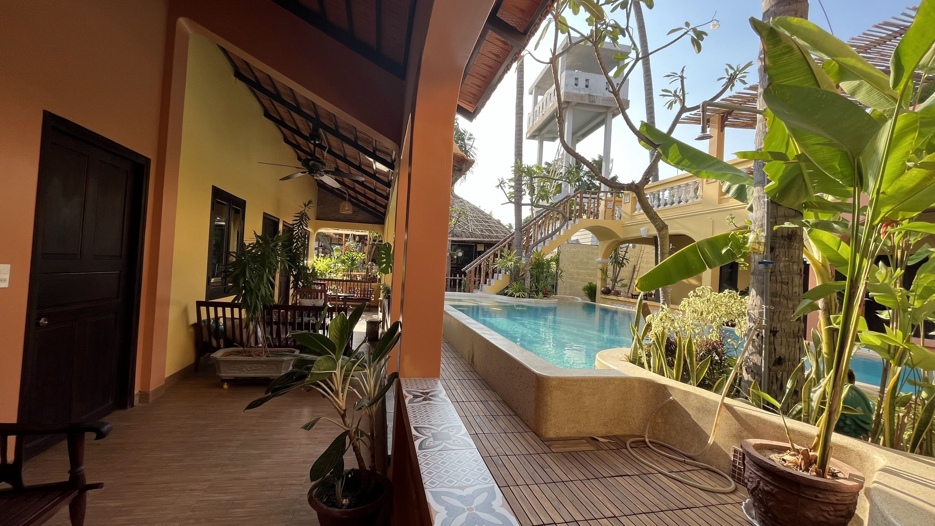 Zona de piscina con terraza de madera, plantas en macetas y un edificio con balcón que da a la piscina.