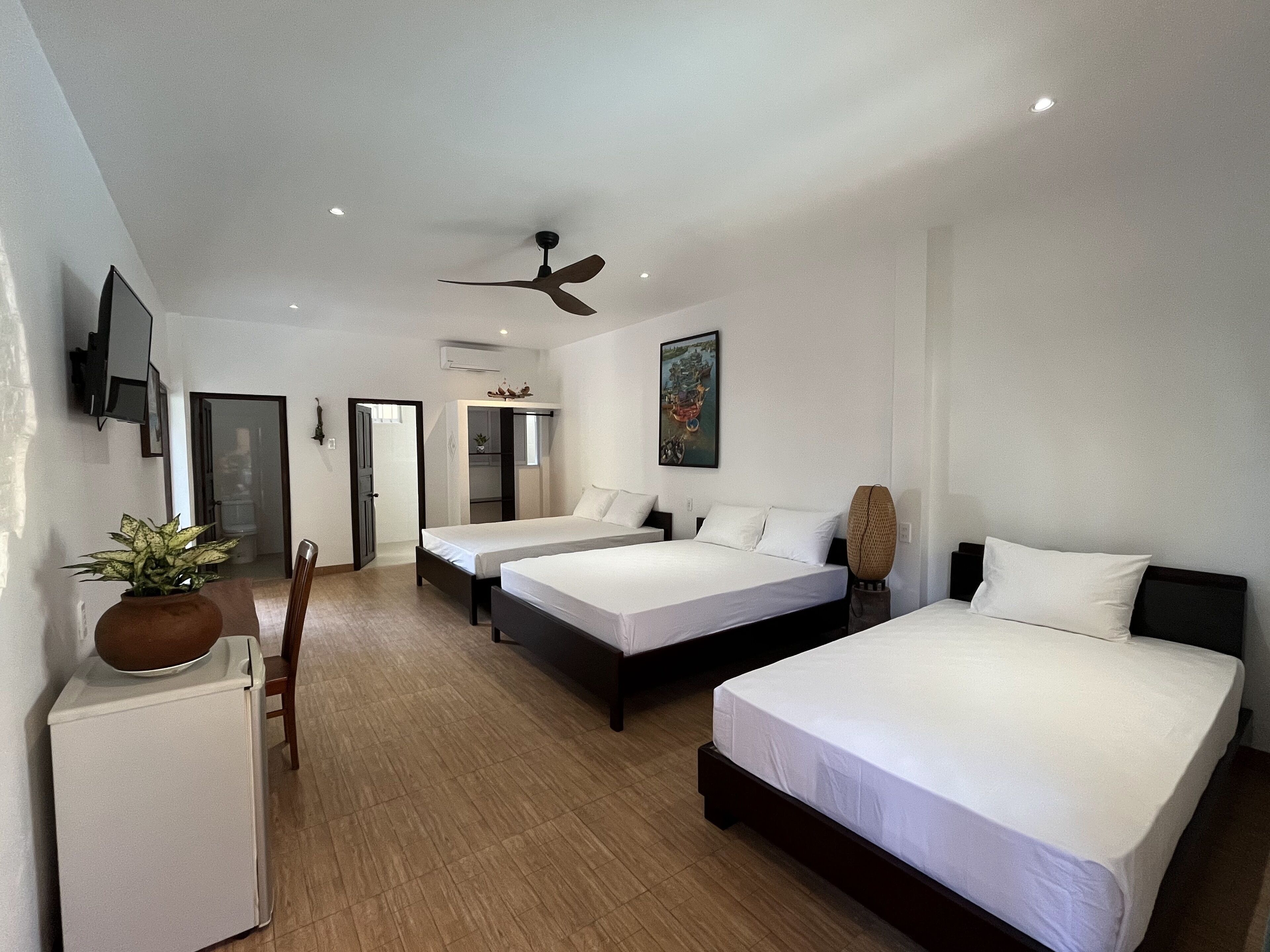 A hotel room with two single beds, a large bed, a ceiling fan, a TV mounted on the wall, and a small refrigerator.
