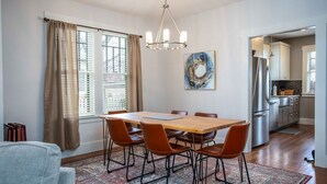 Een eetgedeelte met een houten tafel en zes stoelen, een kroonluchter, een groot abstract schilderij en een keuken op de achtergrond.