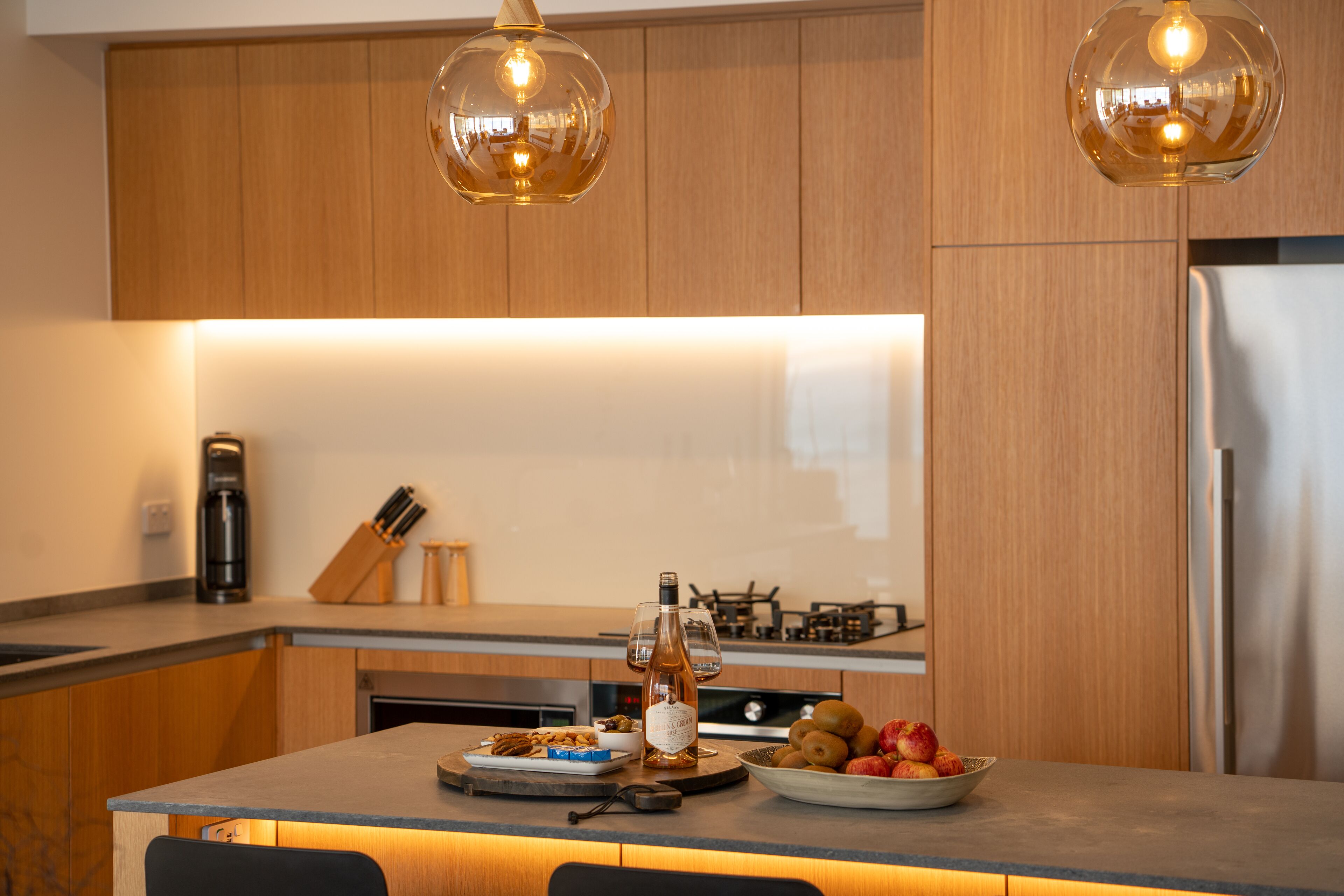 A modern kitchen with wooden cabinets, a stainless steel refrigerator, a gas stove, and a bowl of fruit on the counter.