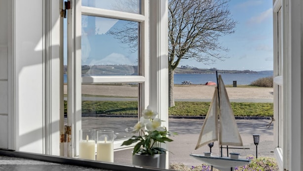 Vista des de l'interior d'una habitació per una finestra, amb una platja, una taula de pícnic, una maqueta de veler i un arbre.