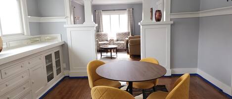 A dining area with a round table and yellow chairs, a kitchen with white cabinets, and a view of a living room with patterned sofas.