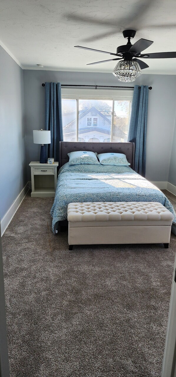 A bedroom with a bed, a nightstand, a lamp, a ceiling fan, and a window with curtains.
