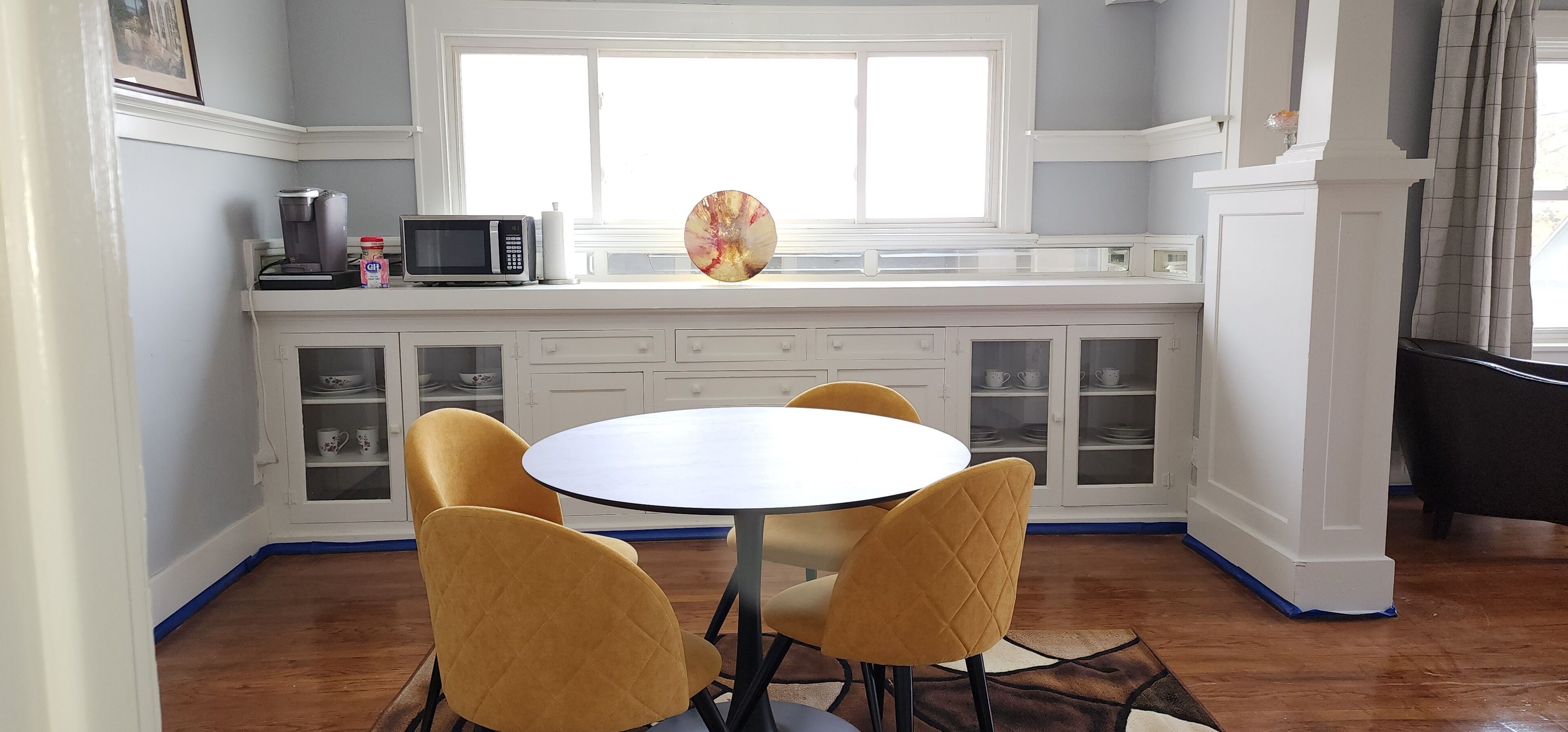 Una cocina con armarios blancos, un microondas y una cafetera sobre la encimera. Hay una mesa redonda con cuatro sillas frente a la cocina.
