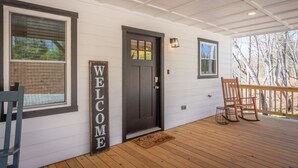 Una terraza de madera con una puerta oscura, un letrero que dice 'WELCOME' y dos mecedoras.