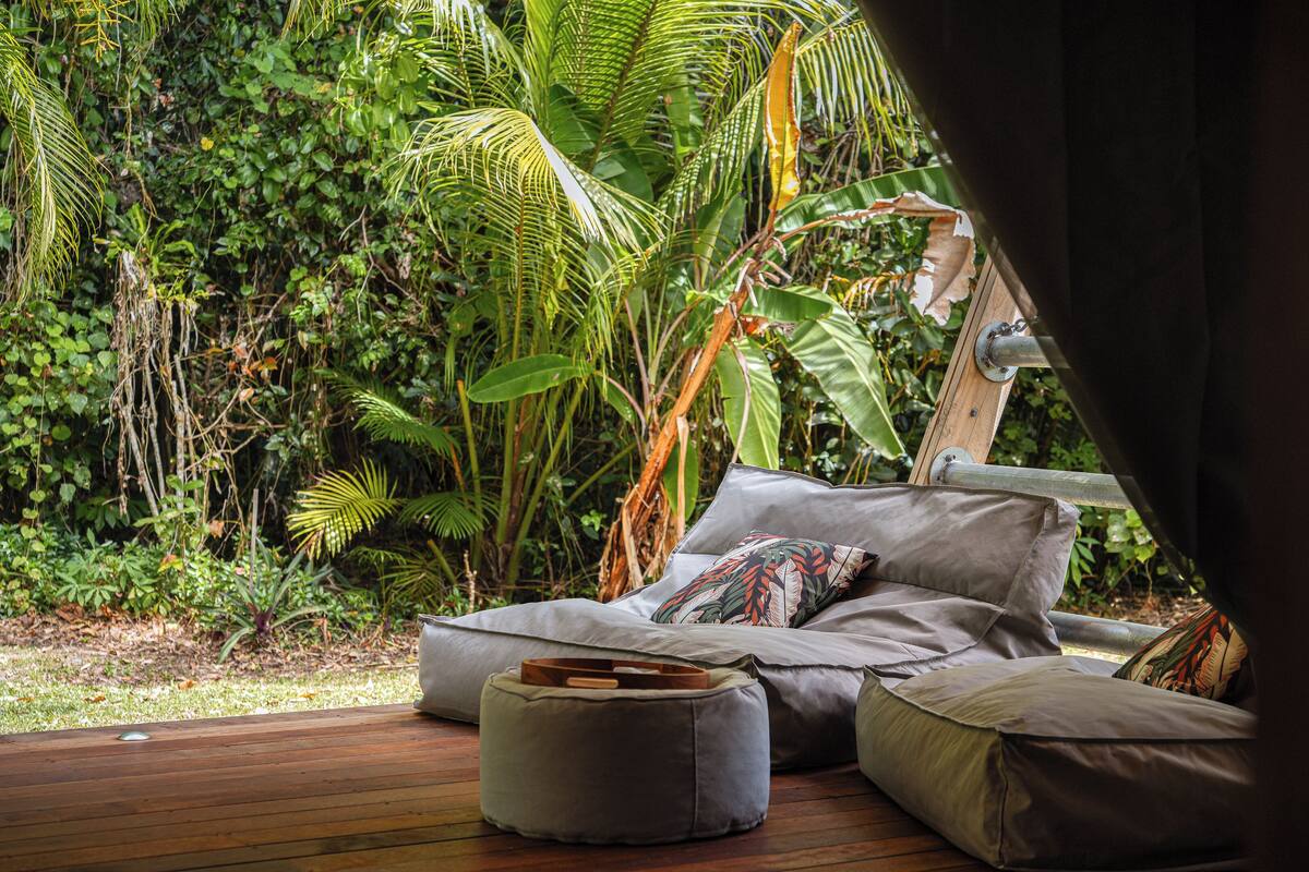 An outdoor seating area with cushions and a wooden deck, surrounded by lush greenery.