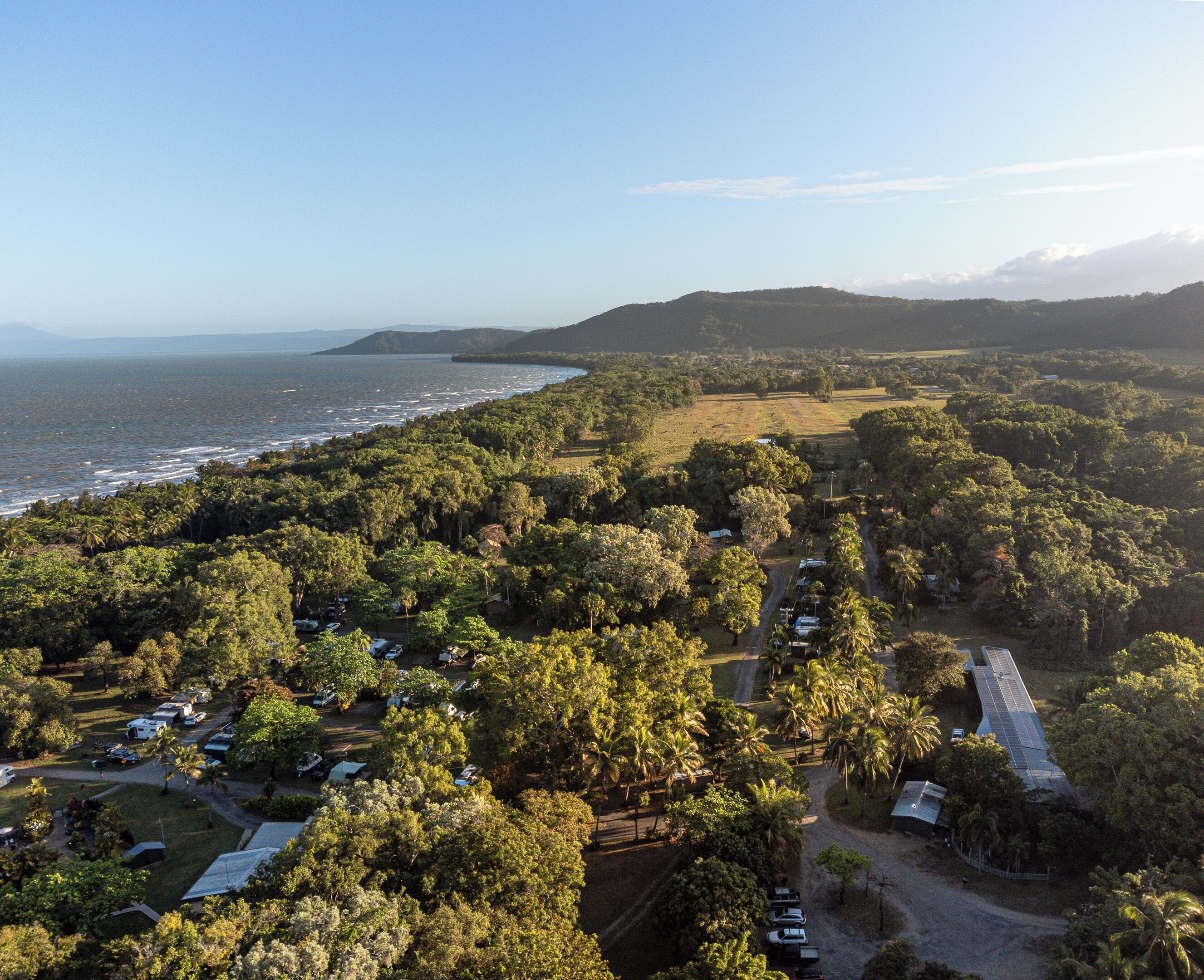 An aerial view of a coastal area with a dense forest, a beach, and a road leading to a small settlement.
