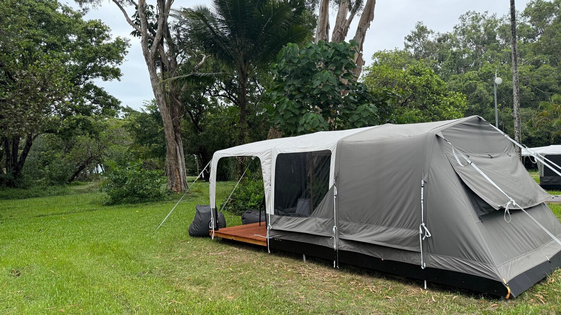 Tienda individual con toldo impermeable, montada en un campo cubierto de hierba con árboles al fondo.