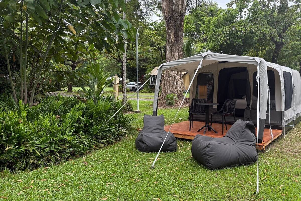 A camping setup with a tent, a table, chairs, and bean bags on a grassy area surrounded by trees.