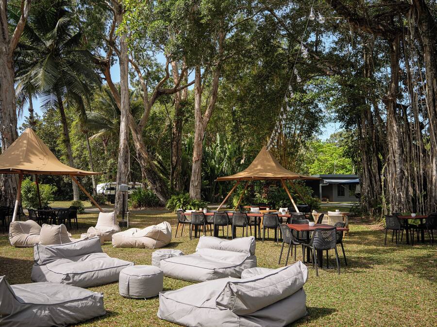 Un espace extérieur aménagé avec des tables et des chaises sous un auvent, entouré de grands arbres et d’une pelouse verdoyante.