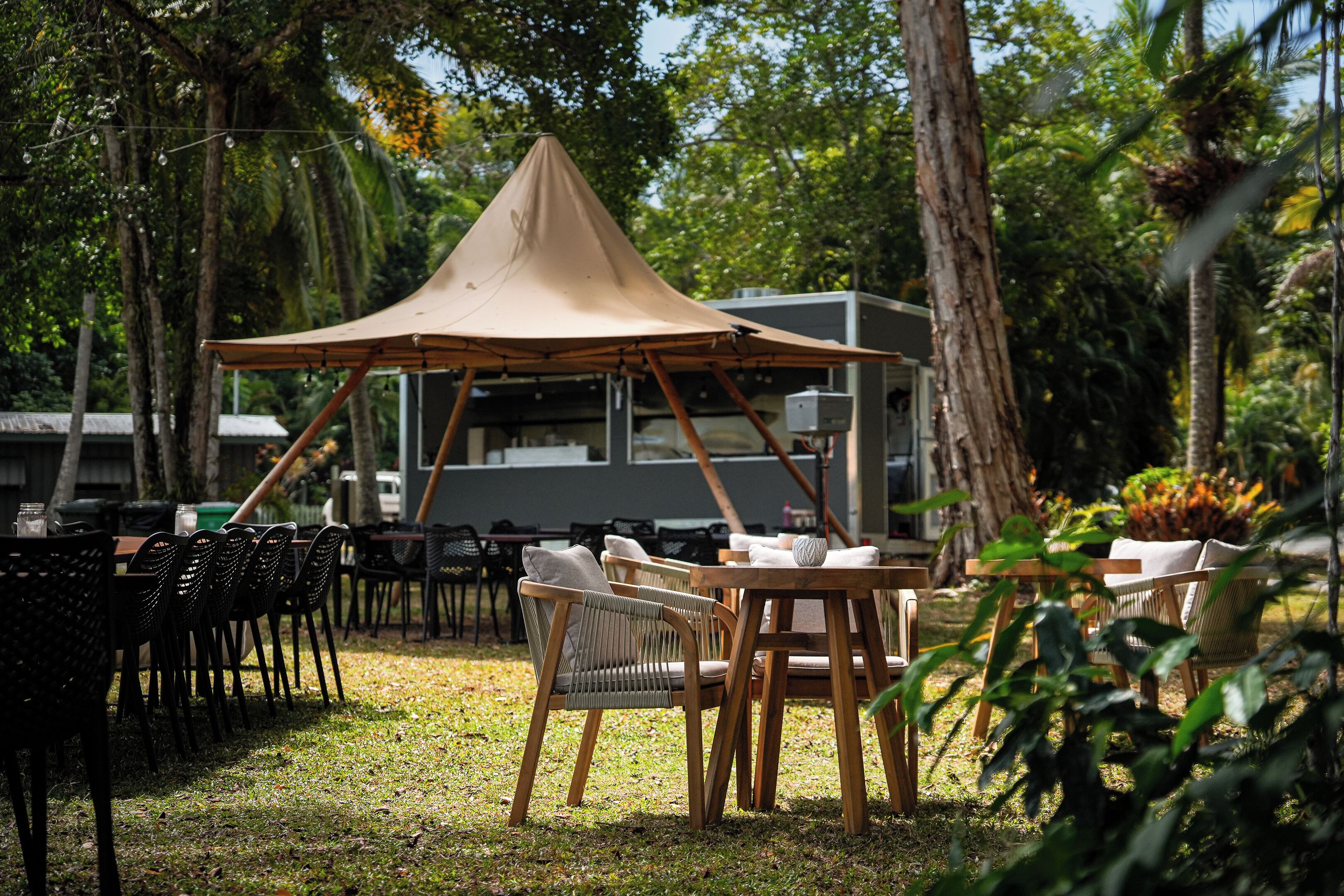 Un espacio de comedor al aire libre con mesas y sillas, un dosel grande, y un edificio al fondo.