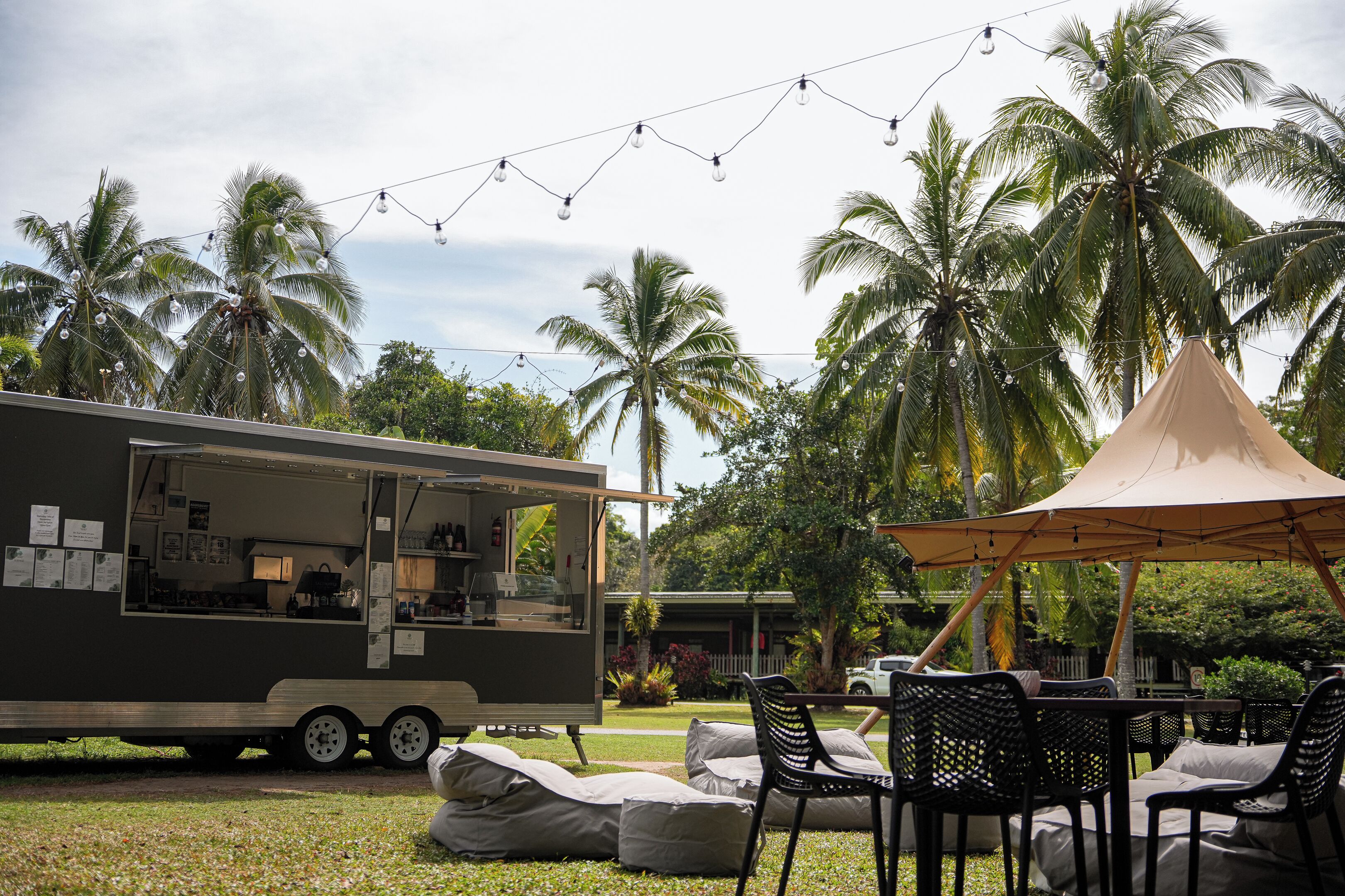 Un camion-restaurant équipé d’une cuisine, avec un espace extérieur aménagé avec des chaises et des coussins, et des palmiers en arrière-plan.