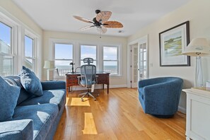 Ein Zimmer mit Holzboden, einem Schreibtisch mit Stuhl, einem blauen Sofa und einem Deckenventilator.