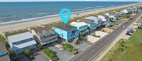 A coastal residential area with houses along a street, a beach in the background, and a parking lot with cars.
