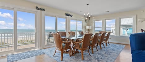 A dining room with a view of the beach, featuring a large table with chairs, a chandelier, and a view of the ocean.