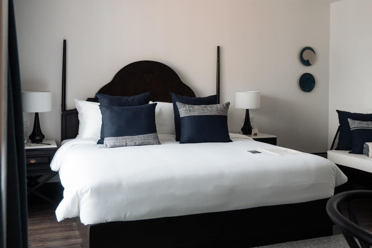 Quarto de hotel com cama grande, cabeceira de madeira escura, roupa de cama branca, duas mesas de cabeceira com abajures, um relógio de parede redondo e um tapete cinza.