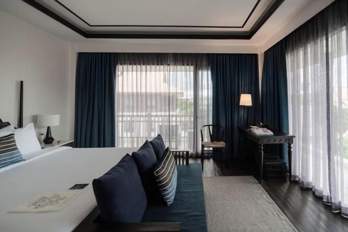 A hotel room with a large bed, a desk with a lamp, a chair, and a view of the outdoors through sheer curtains.
