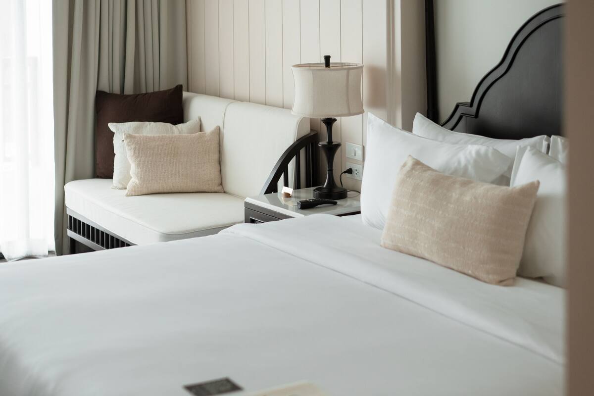 Quarto de hotel com cama bem arrumada, mesa de cabeceira com luminária e janela com cortinas.