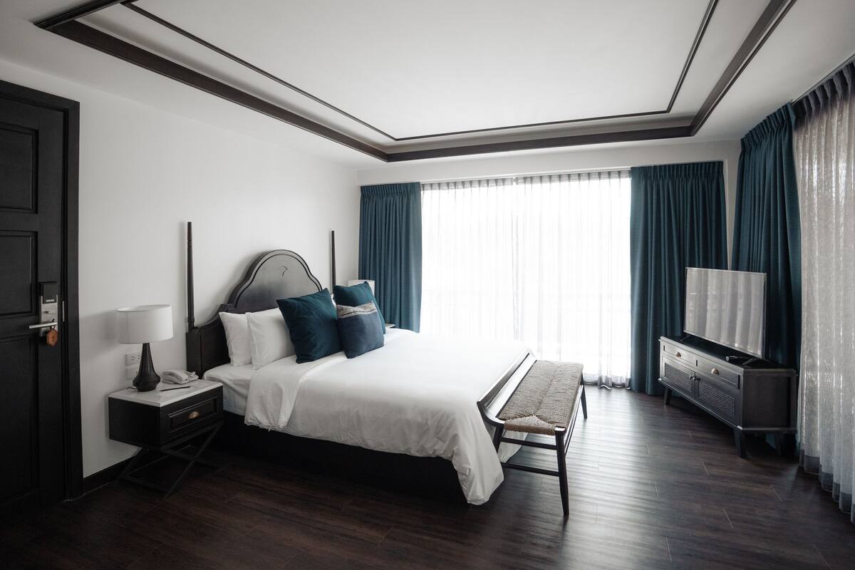 Quarto de hotel moderno com uma cama grande, mesa de cabeceira com abajur, um banco, televisão e guarda-roupa.