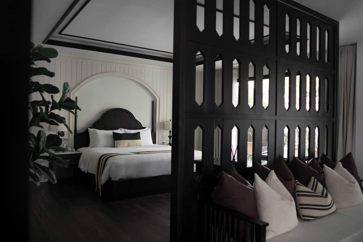 Quarto com cama, mesa de cabeceira, abajur e divisória decorativa com janelas recortadas.