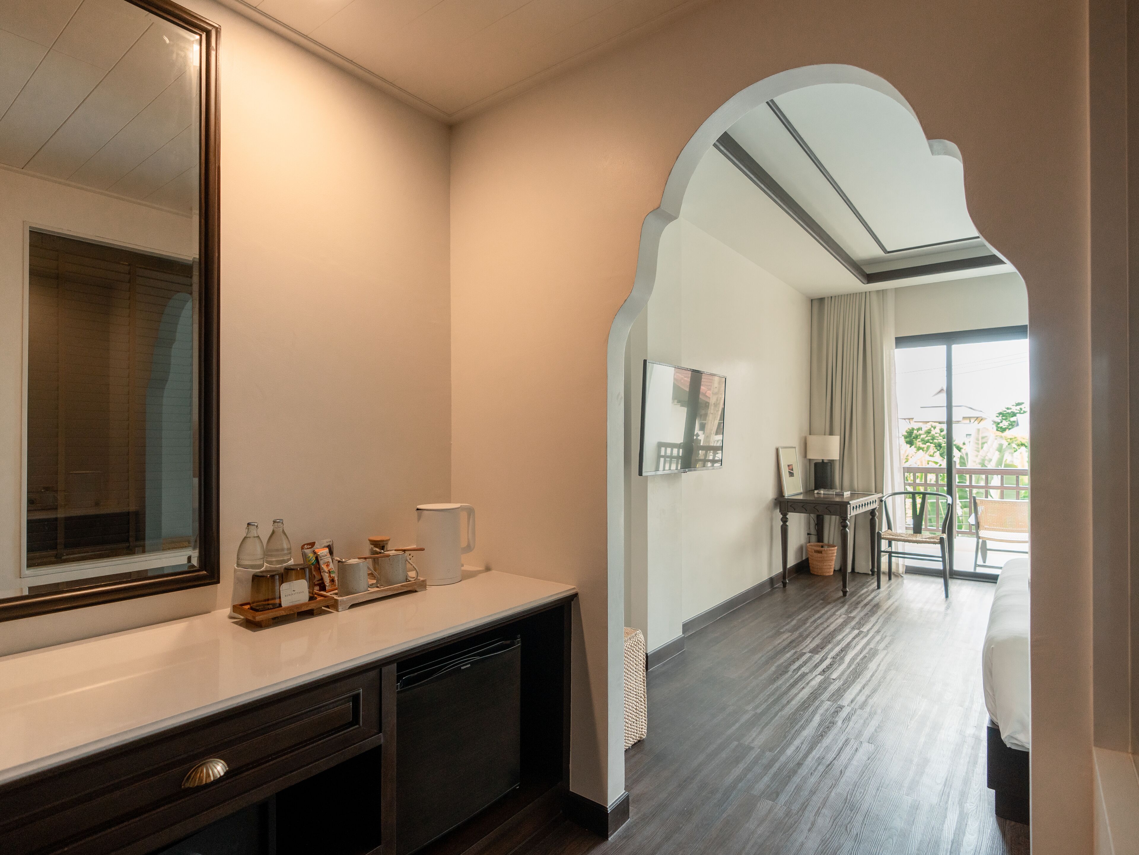 A hotel room with a large mirror, a wooden dresser, a small table with a lamp, and a view of the outdoors through a balcony.