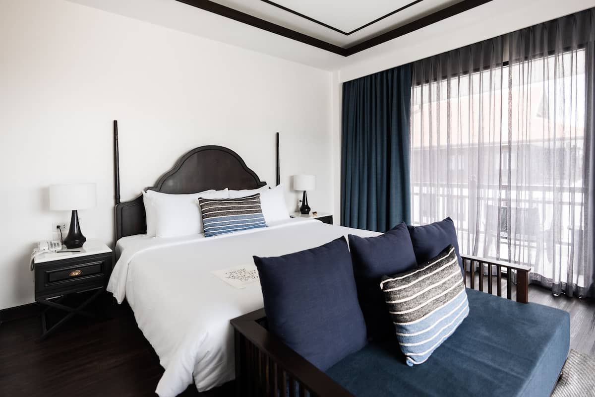 Quarto de hotel com uma cama grande, cabeceira escura, roupa de cama branca e vista para uma varanda através de cortinas leves.