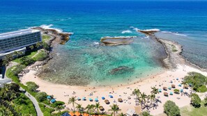 Uma praia com águas turquesa cristalinas, palmeiras e um edifício moderno com fachada de vidro.