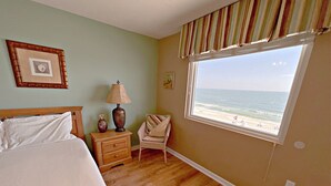 A bedroom with a large bed, a nightstand with a lamp, a chair, a window with a beach view, and a framed picture on the wall. - 👣 Come get your toes in the sand ~~~ and a cold drink in your hand! 🍹 (Gulf Shores)