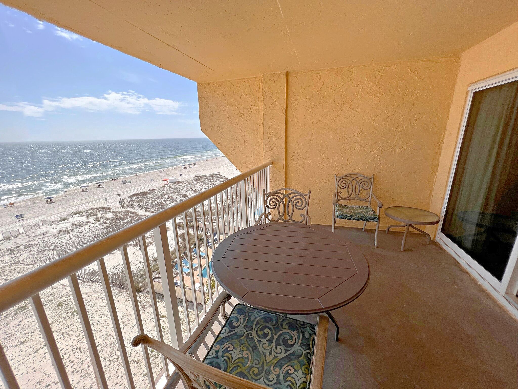 A balcony with a view of the beach, a round table, two chairs, and a glass door.