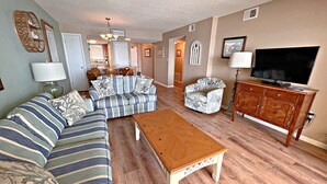 A living room with a sectional sofa, a patterned armchair, a wooden coffee table, and a television on a wooden cabinet. - 👣 Come get your toes in the sand ~~~ and a cold drink in your hand! 🍹 (Gulf Shores)