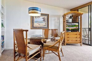 Uma área de refeições com uma mesa de vidro, cadeiras de vime, um armário de madeira com uma televisão e um quadro na parede.