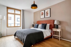 A modern bedroom with a wooden floor, a large bed with a red headboard, two bedside tables with lamps, and a window with curtains.
