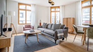 A modern living room with a sofa, coffee table, and a TV on a stand. There are windows with curtains, a lamp, and a cabinet.