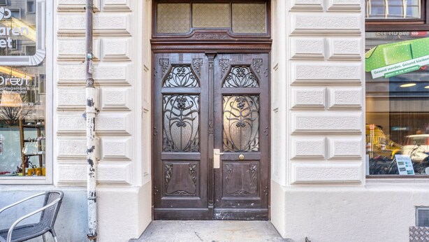 A traditional wooden double door with intricate metalwork, set in a building with a white facade and a small step leading to the entrance.