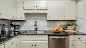 Una cocina con armarios blancos, un lavamanos de acero inoxidable, una cafetera y un lavavajillas.