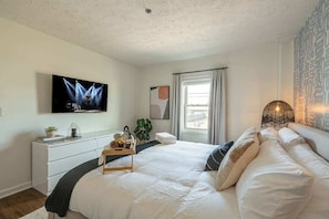 Une chambre à coucher avec un lit, un téléviseur fixé au mur, une table de chevet avec un plateau, une fenêtre avec des rideaux et un mur orné d’un motif.