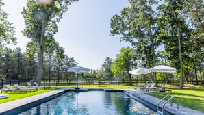 Une piscine rectangulaire à l’eau d’un bleu limpide, entourée d’une pelouse agrémentée de plusieurs parasols blancs et de chaises longues.