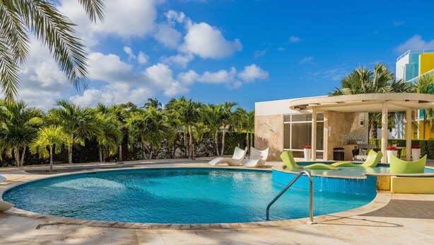 A swimming pool with a curved design, surrounded by palm trees and a covered seating area with chairs.