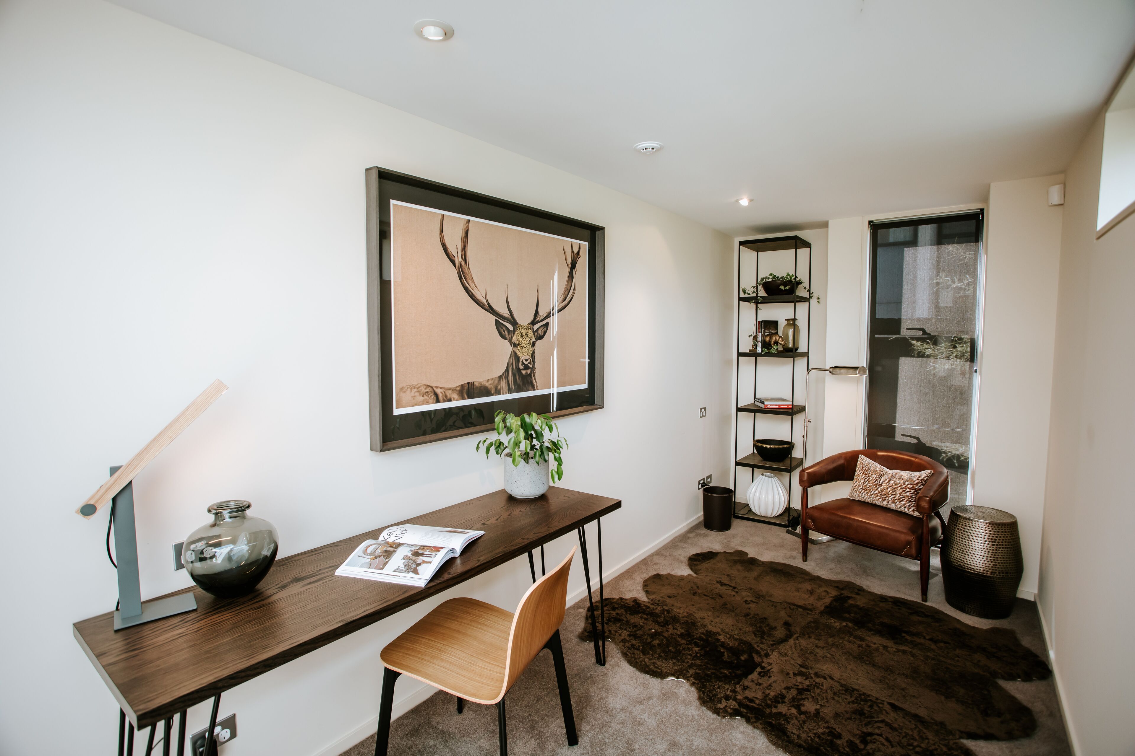 A modern living room with a wooden desk, a leather chair, a large framed deer painting, a potted plant, and a shaggy brown rug.