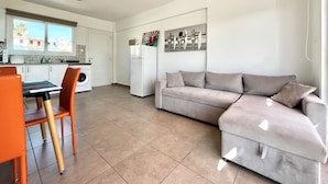 A modern kitchen with white cabinets, a dining table with orange chairs, and a grey sofa. - STAY Symphony Apartment (Protaras)