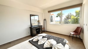 A hotel room with a bed, a desk with a mirror, a chair, and a large window with a view of trees and the sea. - STAY Symphony Apartment (Protaras)