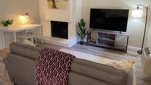 A living room with a white brick fireplace, a flat-screen TV, a sofa, a desk with a chair, and a lamp. - Cozy 1K & 1Q / 1b, 5 minutes walk to downtown (Lafayette)