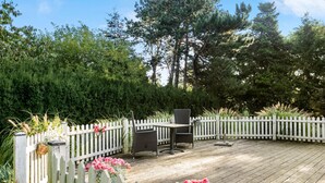 Eine Holzterrasse mit weißem Zierzaun, umgeben von dichtem Grünwerk und hohen Bäumen.