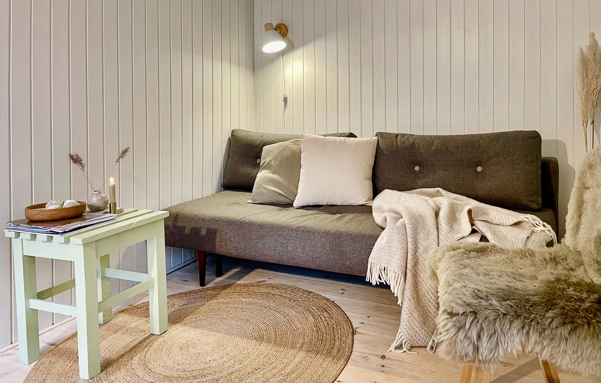 Ein gemütlicher Raum mit einem grünen Sofa, einem kleinen Tisch mit Schale und Kerzen, einem gewebten Teppich und einem flauschigen Sessel.