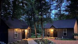 Dos cabañas con paredes de madera y techos oscuros, unidas por un sendero de madera, rodeadas de árboles.