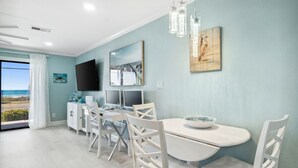 A modern dining area with a white table and chairs, a flat-screen TV, and artwork on the wall. - LAGUNA DUNES! AMAZING VIEWS! GROUND FLOOR! (Panama City Beach)
