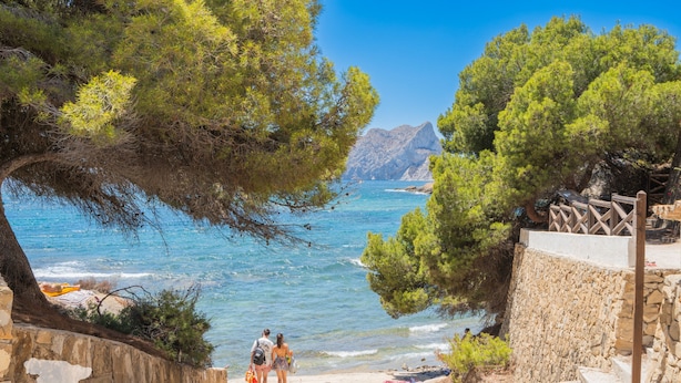 Eine Steintreppe führt zu einem Strand mit klarem, blauem Wasser und felsigen Bergen im Hintergrund.