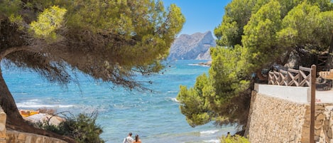 Un escalier en pierre mène à une plage aux eaux d’un bleu limpide, avec des montagnes rocheuses en toile de fond.