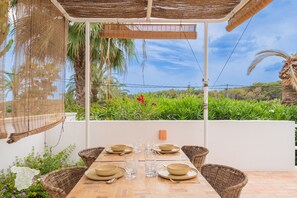 Ein Essbereich mit Tisch für vier Personen, umgeben von Korbstühlen, unter einer Pergola mit Blick auf einen üppigen Garten und Palmen.